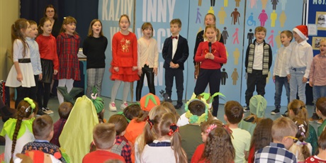 Powiększ grafikę: Dziewczynki i chłopcy z klasy I stoją na scenie i spiewają piosenkę. Przed nimi stoi solistka z mikrofonem w czerwonym sweterku.